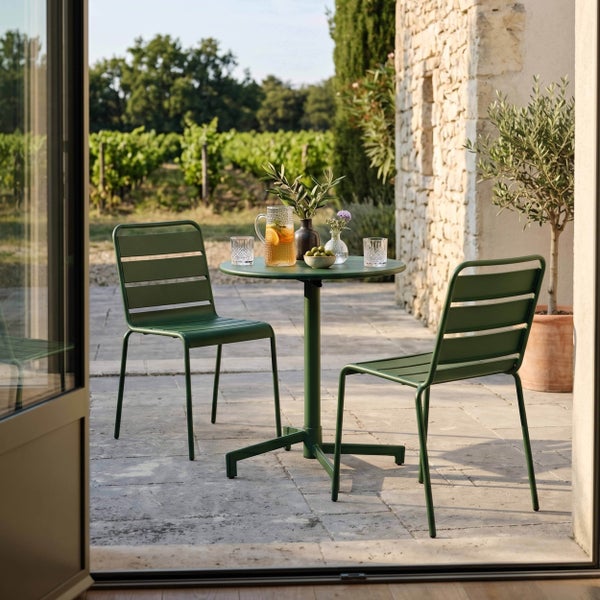 Grünes Garten-Bistro-Set aus Metall mit zwei Stühlen und einem runden Tisch auf einer hellen Steinterrasse vor einem Weinberg.