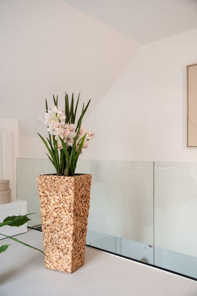 Orchidee mit Blüten in einem hohen, geflochtenen Pflanzgefäß aus Naturmaterialien in einem hellen, modernen Raum mit Glasgeländer.