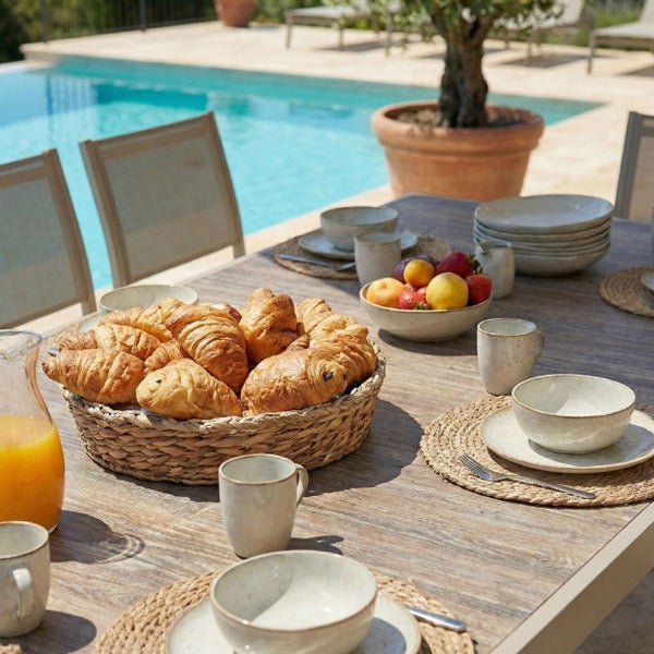 Gedeckter Gartentisch aus Holz mit Keramikgeschirr, Gebäckkorb und Obstschale im hellen Außenbereich neben einem Pool.