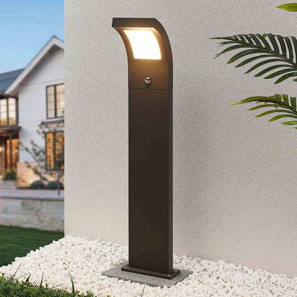 Anthrazitfarbene LED-Sockelleuchte mit Bewegungsmelder im modernen Design vor einer Hauswand auf weißen Kieselsteinen.