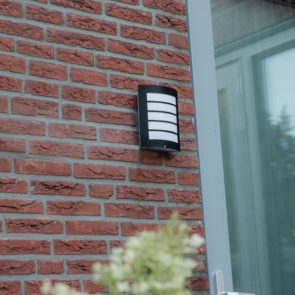 Moderne schwarze Außenwandleuchte mit horizontalen Lamellen an einer Ziegelwand neben einer Tür.