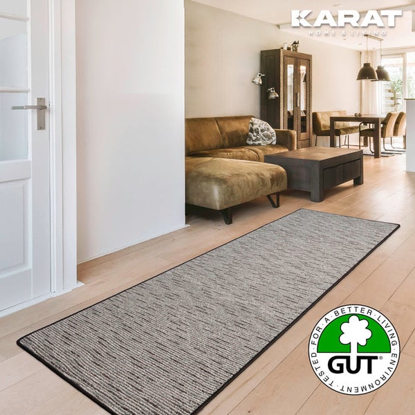 Grauer strukturierter Teppichläufer mit schwarzem Rand im hellen Wohnzimmer. Geprüft auf Umweltverträglichkeit. Karat Home and Living Logo.