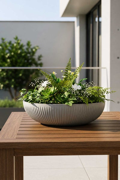 Graue, geriffelte Pflanzschale auf einem Holztisch, bepflanzt mit Sukkulenten, Farnen und weißen Blumen im modernen Außenbereich.