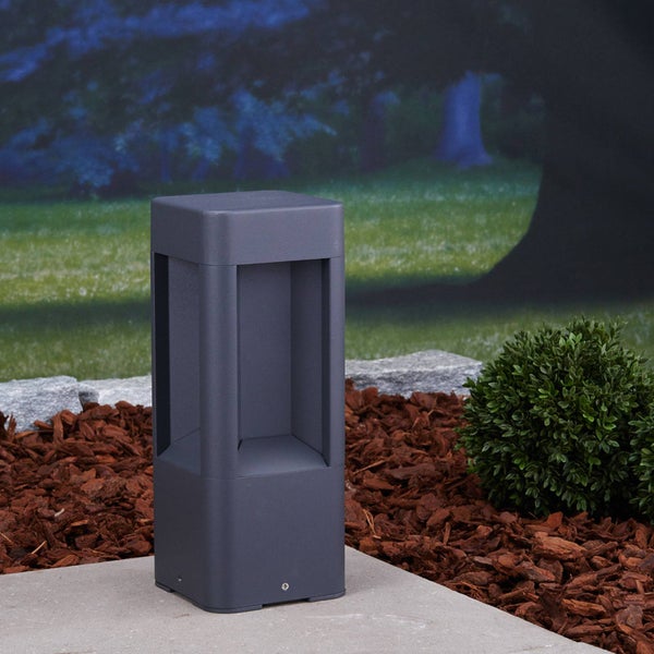 Moderne, quadratische Pollerleuchte in Anthrazit auf einer Betonplatte im Garten, umgeben von Rindenmulch und einem kleinen Strauch bei Abenddämmerung.