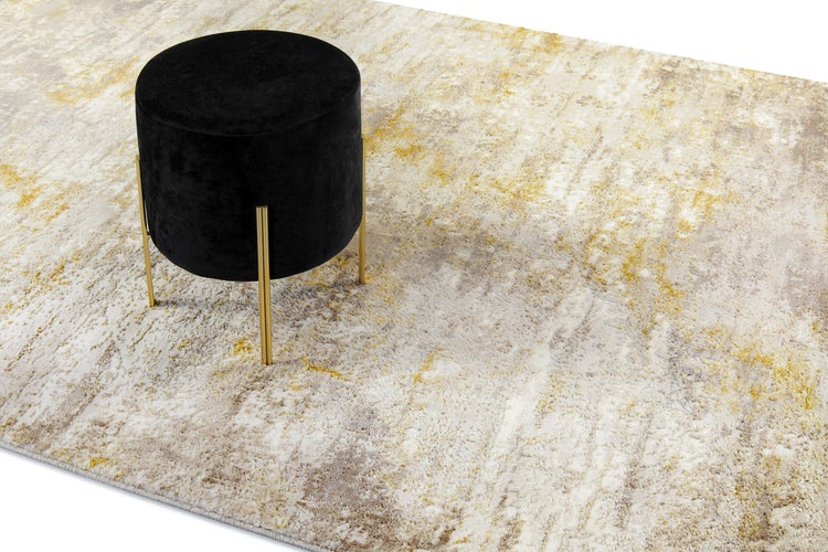 Schwarzer Samthocker mit goldenen Metallbeinen auf einem gemusterten Teppich mit abstraktem Design in Beige und Ocker.