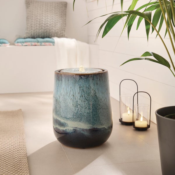 Keramik-Zimmerbrunnen in Form eines runden Gefäßes mit blau-grüner Glasur, im Hintergrund Kerzenleuchter und eine Pflanze.