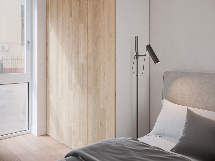Helles Schlafzimmer mit Holzschrank, Bett mit grauem Kopfteil und einer schmalen, schwarzen Stehleuchte neben einem großen Fenster.