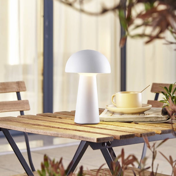 Weiße LED-Tischleuchte in Pilzform auf einem Holztisch im Außenbereich mit einer Tasse und einer Zeitung.