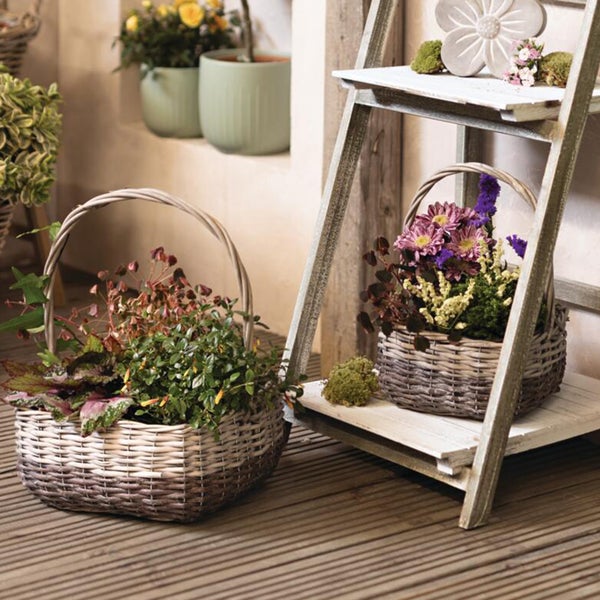 Zwei Weidenkörbe mit Griffen, bepflanzt mit Blumen und Grünpflanzen, auf einem Holzdeck und einer dekorativen Holzleiter.