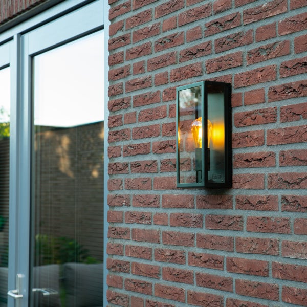 Moderne rechteckige Außenwandleuchte aus schwarzem Metall und Glas mit warm leuchtendem Leuchtmittel an einer roten Backsteinwand.