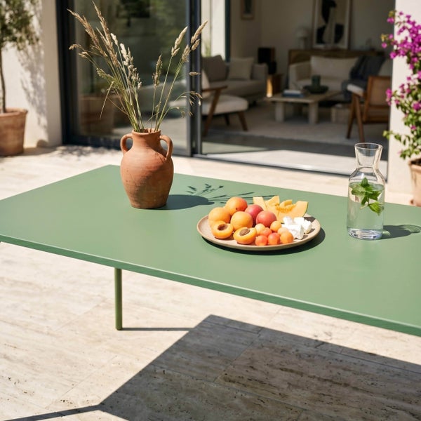 Moderner, grüner Gartentisch aus Metall auf einer hellen Steinterrasse, dekoriert mit einer Terrakotta-Vase, Früchten und einer Wasserkaraffe.