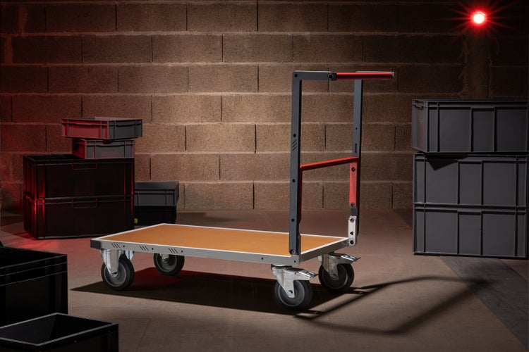 Transportwagen mit Ladefläche und vier Rädern vor einer Ziegelwand und mehreren Lagerboxen