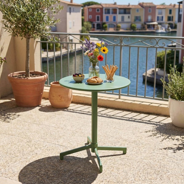 Grüner, runder Bistrotisch aus Metall auf einer sonnigen Terrasse am Wasser, gedeckt mit Blumen und Snacks.