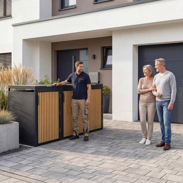 Moderne Mülltonnenbox aus Metall und Holz vor einem Wohnhaus, präsentiert von einem Fachberater auf einer gepflasterten Einfahrt.