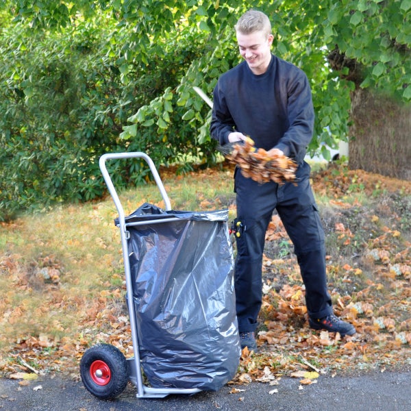 Person füllt Herbstlaub in einen Abfallsack auf einem fahrbaren Gartenwagen aus Metall im Außenbereich.