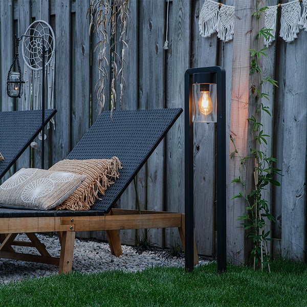 Schwarze Solar-Stehleuchte mit Vintage-Leuchtmittel in einem Garten neben einer Sonnenliege aus Holz und einem Holzzaun.