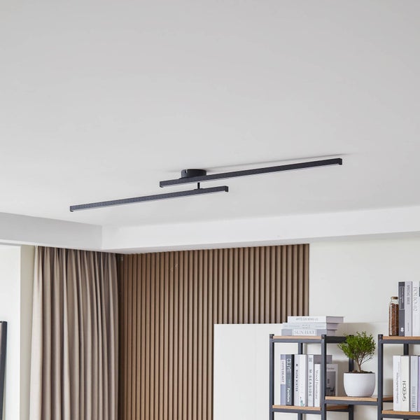 Moderne schwarze Deckenleuchte mit zwei langen, schmalen Stäben an einer weißen Decke über einem Bücherregal vor einer Holzlamellenwand.