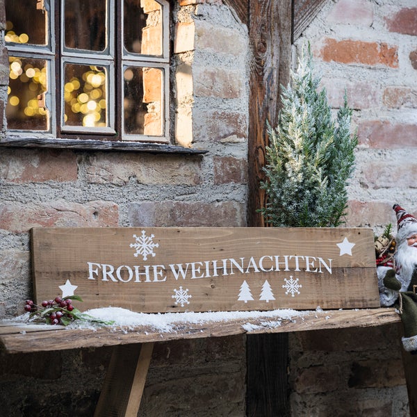 Dekorative Holzbank mit der Aufschrift 'Frohe Weihnachten', dekoriert mit Kunstschnee, Beeren, einer Miniaturtanne und einer Weihnachtsmannfigur vor einer Backsteinmauer.
