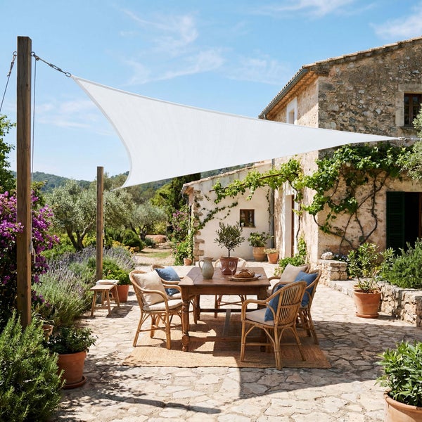 Dreieckiges Sonnensegel über einer Terrasse mit Holztisch und Rattanstühlen vor einem Steinhaus in mediterraner Gartenumgebung.