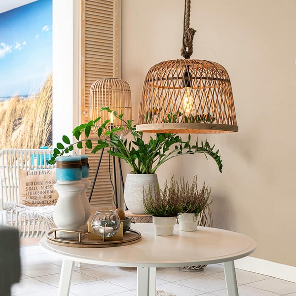 Dekoratives Interieur mit hängender Korblampe, Tischlampe, Pflanzen und Kerzen auf einem runden Tisch.