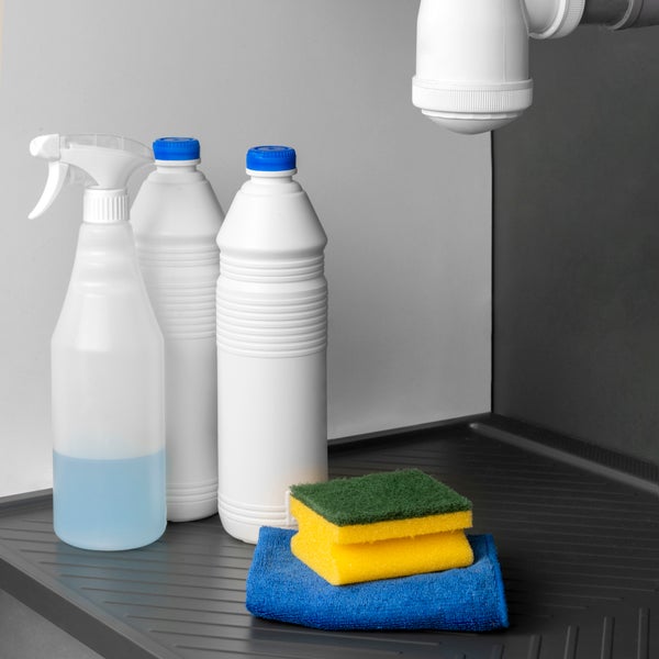 Reinigungsutensilien wie Sprühflasche, Putzmittel, Schwamm und Tuch auf einer schwarzen Unterschrankmatte unter einem Siphon.