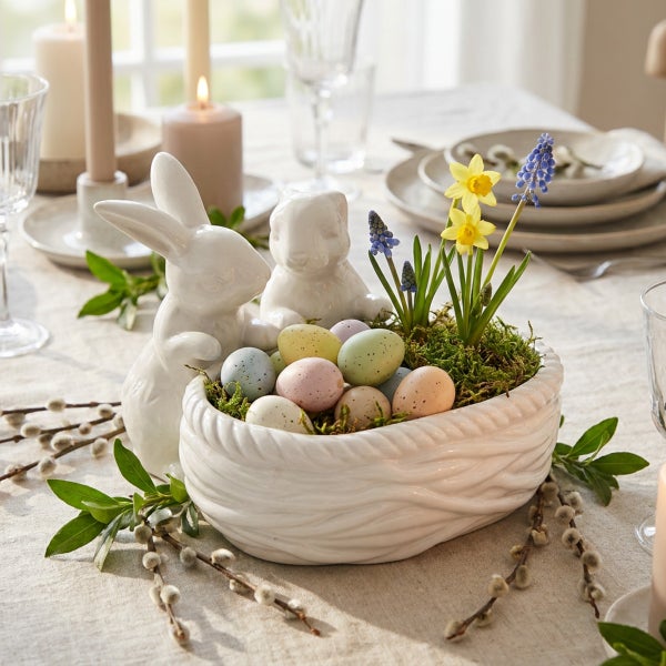 Osterdekoration mit zwei Keramikhasen und einem geflochtenen Keramikkorb gefüllt mit Ostereiern und Frühlingsblumen auf einem gedeckten Tisch.