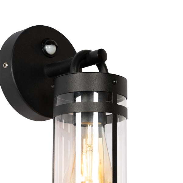 Außenwandleuchte mit Bewegungsmelder, schwarzes Metallgehäuse, zylindrischer Glasschirm mit sichtbarem Leuchtmittel.