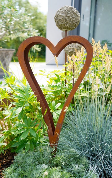 Gartendekoration Herz aus Metall in Edelrost Optik platziert in einem bepflanzten Gartenbeet.