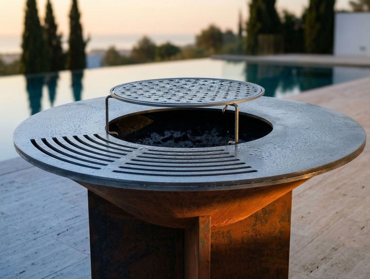 Moderner Feuerstellen-Grill aus Metall mit runder Grillplatte und Aufsatzgitter auf einer Terrasse an einem Pool bei Abenddämmerung.