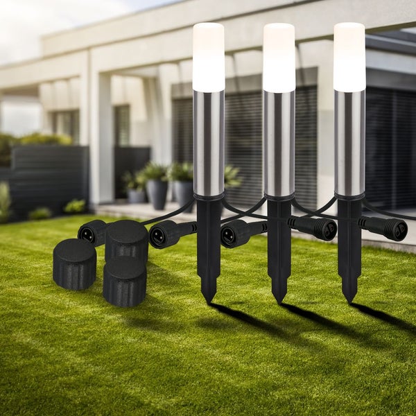 Dreiteiliges Set Gartenleuchten aus Edelstahl mit Erdspießen, warmweißem Licht und Kabelverbindung auf einer Rasenfläche vor einem Haus.