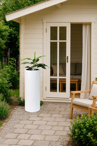 Hoher, weißer zylindrischer Pflanzkübel mit grüner Pflanze auf gepflastertem Gartenweg vor einem hellen Gartenhaus aus Holz.