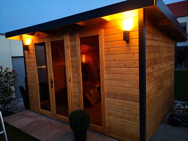 Modernes Gartenhaus aus Holz mit Flachdach, Doppelflügeltür aus Glas und Außenbeleuchtung am Abend.