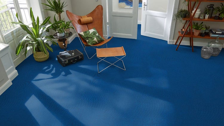 Heller Wohnraum mit blauen, strukturierten Teppichfliesen, einem braunen Ledersessel, Zimmerpflanzen und einer weißen Tür.