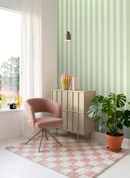Hell gestalteter Raum mit Tapete im vertikalen Kreismuster, rosa Sessel, beigem Sideboard, kariertem Teppich und großer Monstera-Pflanze.