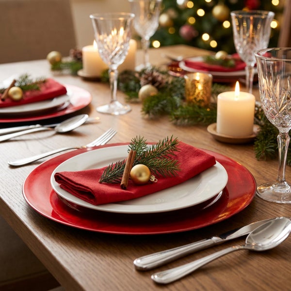 Festlich gedeckter Esstisch mit weihnachtlicher Dekoration, Tellern, Besteck, Gläsern, Kerzen und Tannenzweigen auf einer Holzoberfläche.