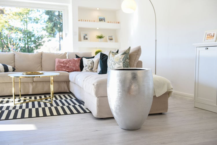 Großer, silberfarbener Pflanzkübel in einem hellen Wohnzimmer mit beigem Sofa und Couchtisch. Modernes Design und strukturierte Oberfläche.