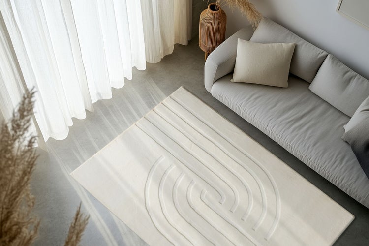 Wohnzimmer mit modernem Teppich mit geometrischem Reliefmuster auf einem Betonboden, neben einem Sofa und Vorhängen in einem hellen Raum.