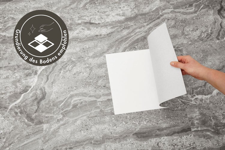 Hand zieht Schutzfolie von selbstklebendem Vinylboden in Steinoptik ab, mit Hinweis auf Grundierung des Bodens.