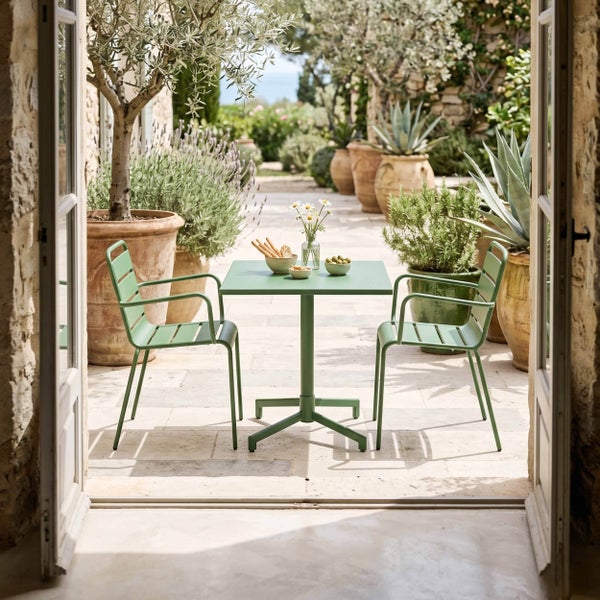 Bistro-Set aus grünem Metall mit Tisch und zwei Stühlen auf einer hellen Steinterrasse mit Lavendel und Olivenbäumen in Terrakottatöpfen.