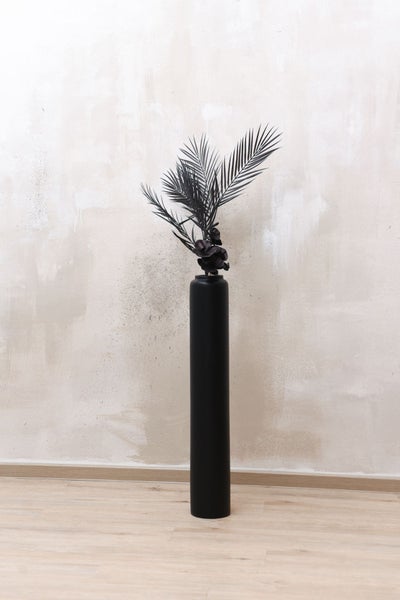 Hohe, zylindrische Bodenvase in Schwarz mit schwarzen künstlichen Palmzweigen und Blüten auf hellem Boden.