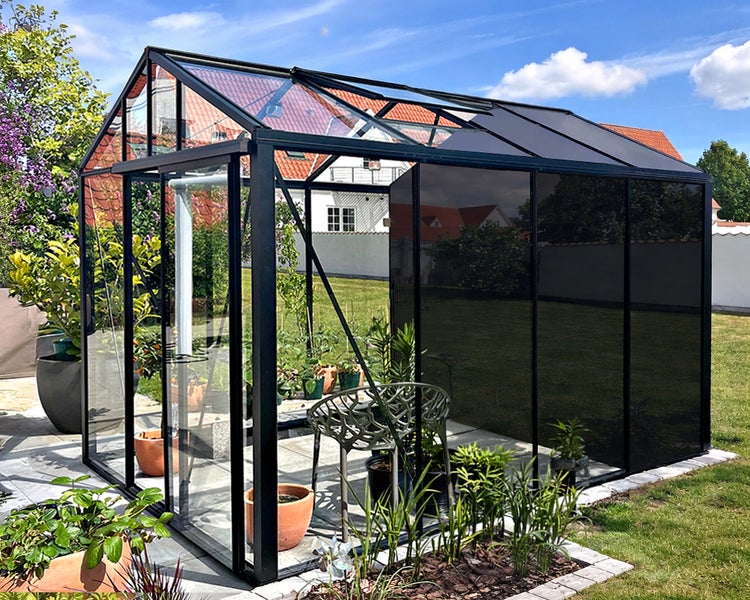 Modernes Gewächshaus mit schwarzem Aluminiumrahmen, klaren und getönten Scheiben, eingerichtet mit Pflanzen und einem Stuhl im hellen Garten.