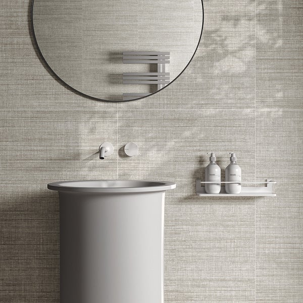 Modernes Badezimmer mit rundem Wandspiegel, Säulenwaschbecken, Wandarmatur und Wandablage mit Seifenspendern vor texturierten Fliesen.