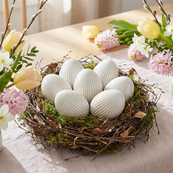 Osternest mit keramischen Ostereiern mit Rillenmuster, umgeben von Frühlingsblumen wie Tulpen, Hyazinthen und Weidenkätzchen auf einem Holztisch.