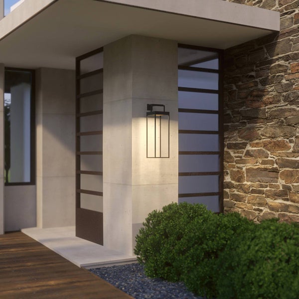 Moderne schwarze Außenwandleuchte an einem Betonpfeiler neben einer Natursteinwand, die warmes Licht auf die Holzterrasse wirft.
