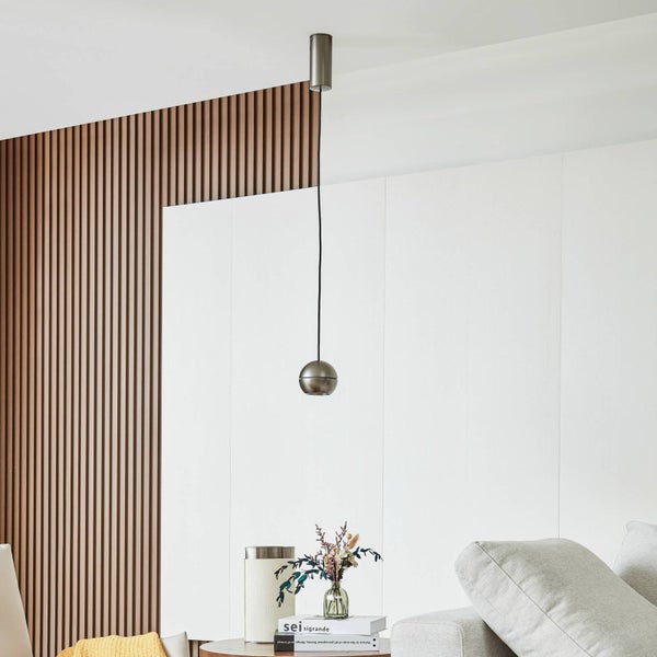 Moderne Pendelleuchte mit kugelförmigem Metallschirm in einem hellen Wohnzimmer mit Holzlamellenwand und Sofa.