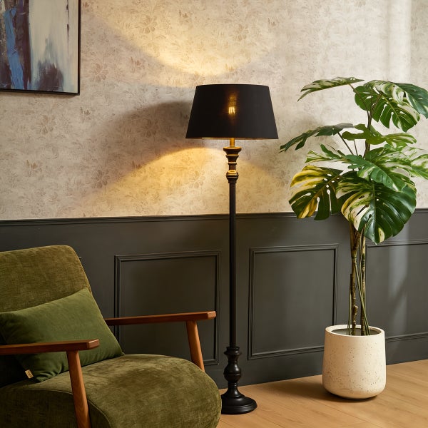 Stehleuchte mit schwarzem Schirm neben einem grünen Sessel und einer Monstera-Pflanze in einem Raum mit gemusterter Tapete und Wandpaneelen.