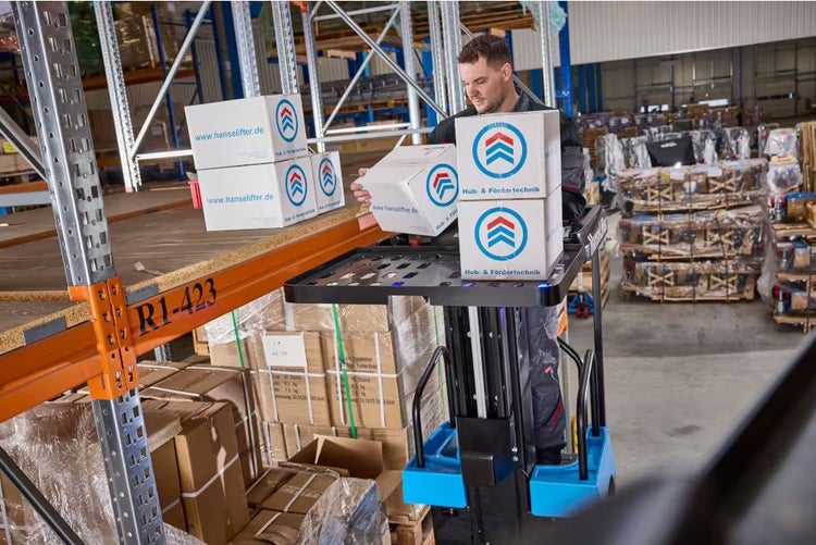 Ein Mann bedient einen elektrischen Kommissionierer, um Kartons mit Hanselifter Logo in ein Lagerregal zu stapeln.
