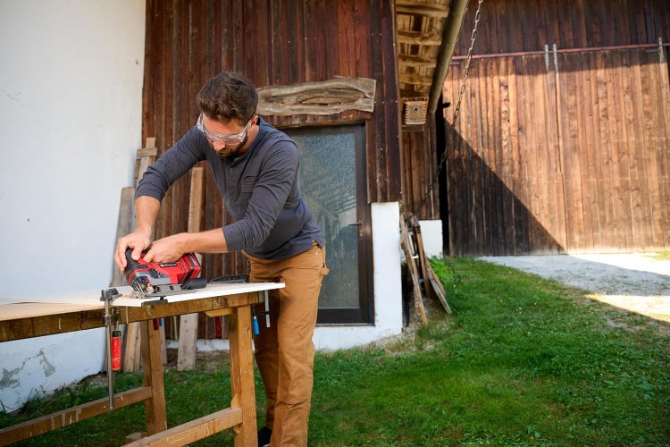 Handwerker mit Schutzbrille sägt Holzplatte mit einer Einhell Stichsäge auf einer Werkbank im Freien vor einer alten Scheune.