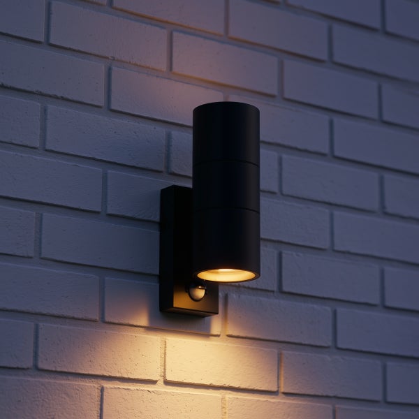 Zylindrische Außenwandleuchte in Schwarz mit Bewegungsmelder, montiert an einer hellen Ziegelwand, warmweißes Licht strahlt nach unten.