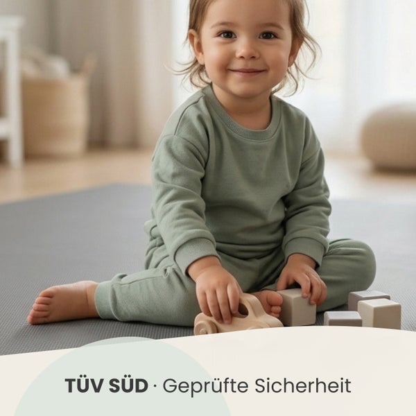 Kleinkind spielt in einem hellen Zimmer auf einer Matte mit Holzspielzeug, TÜV SÜD geprüfte Sicherheit.